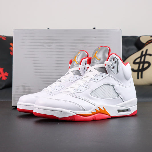 A pair of white Air Jordan 5 Retro sneakers with red and orange accents, positioned in front of their silver box on a dark surface.